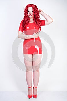 Hot sexual redheaded girl with plus size body wears fashion latex rubber red dress and posing on white studio background alone. ch
