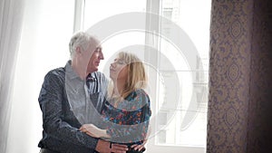 Hot and Sexy Middle-aged Woman Enjoying Hug of Her Elderly Husband Standing near Opened Window inside Their Home. Couple