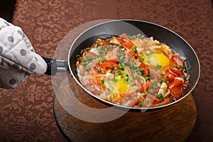 hot shakshuka in a frying pan sprinkled with green onions is holding a potholder.