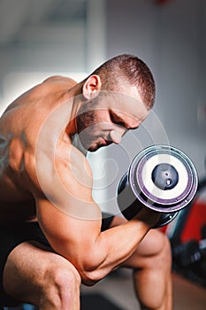 A hot sportsman with a naked torso lifting a dumbbell in a gym on a blurred light background.