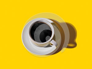 Hot strong black coffee in white elegant cup with saucer over a bright yellow background. Hot morning beverage. Shot with hard