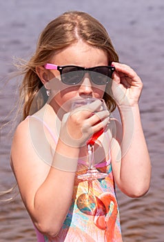 On a hot summer day, a child is relaxing on the lake. A girl on the beach. The girl drinks delicious fresh juice.