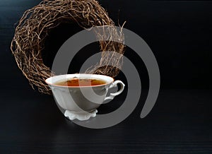 Hot tea cup white porselain with round wicker wreath decoration at black wooden background/Retro