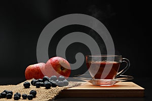 The hot tea fruits in glass cup with smoke and blueberry, apple on the wooden board as black background. Healthy drink concept.