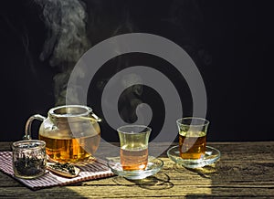 Hot tea in glass teapot and cup with steam on wooden table, black background