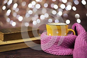 Hot tea, hot chocolate, coffee in yellow cup, wrapped with a pink knitted scarf. Old books. Blurred lights, wooden background.