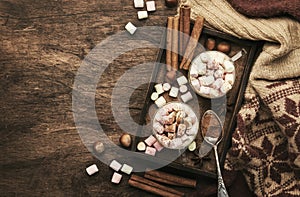 Hot winter or autumn drink with cocoa, chocolate, spices and marshmallows in cups on vintage wooden background with knitted warm