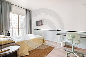 Hotel room with bed for two persons with beige linen. Access to balcony through glass sliding doors. Table against wall