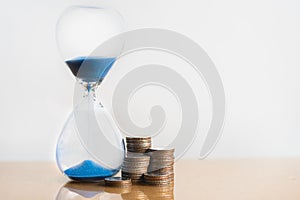 Hourglass blue sand and stack of coins.