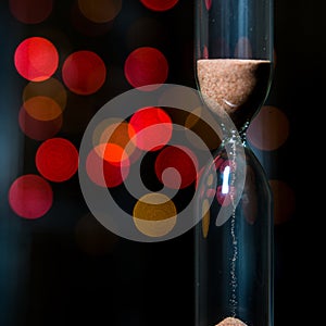 Hourglass in the center. Sand strews down on christmas background, bokeh red christmas lights.