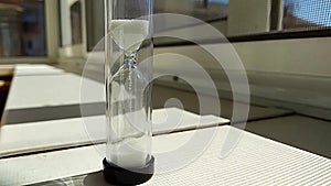 Hourglass with a fleeting white sand inside, stand on a window sill, time lapse