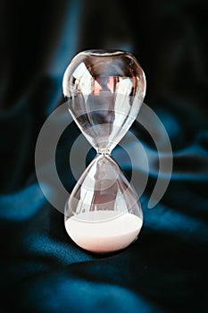 An hourglass with pink sand at the bottom of the clock is on a blue satin background. The concept of stopped time.