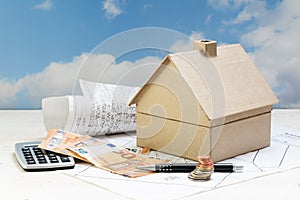 house from cardboard with money and calculator standing on architecture plans against a blue sky with clouds, copy space