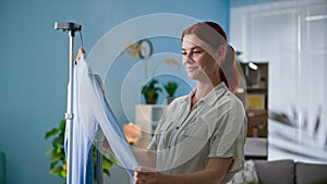 house cleaning, smiling woman using modern iron steams clothes on hanger at home