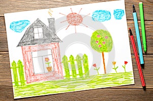 A house, a fence, flowers and an apple tree. Children's drawing on paper. Wooden table with markers