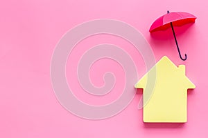 House insurance concept. Toy house defended by umbrella on pink backgound top view copy space