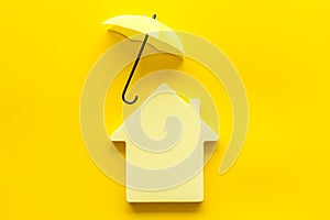 House insurance concept. Toy house defended by umbrella on yellow backgound top view copy space