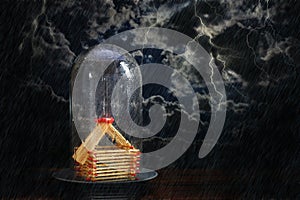House from matches covered under a glass dome against a dark stormy sky with lightning, insurance concept for real estate