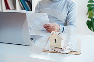 House model with British currency on table and blurred man with docs works on laptop. Housing cost and payments. Real