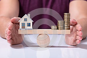 House Model And Money Coins Balancing On A Seesaw