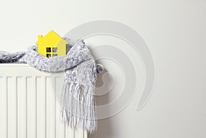 House model wrapped in scarf on radiator, space for text. Winter heating efficiency