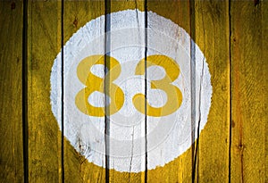 House number or calendar date in white circle on yellow toned wooden door background. Number eighty three 83