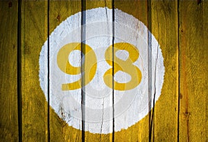 House number or calendar date in white circle on yellow toned wooden door background. Number ninety eight, 98