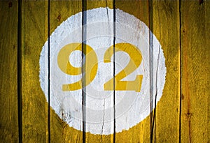 House number or calendar date in white circle on yellow toned wooden door background. Number ninety two 92
