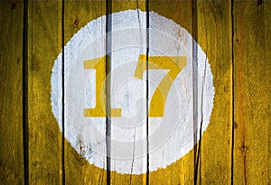 House number or calendar date in white circle on yellow toned wooden door background. Number seventeen 17