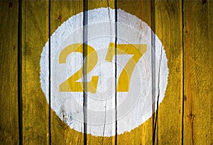 House number or calendar date in white circle on yellow toned wooden door background. Number twenty seven 27