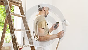 House painter man with mobile phone and paint roller, works the white home wall, a wooden ladder and green window as a background