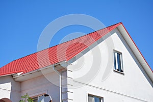 House with plaster walls, attic window, red metal roof and white plastic rain gutter. Roofing construction.