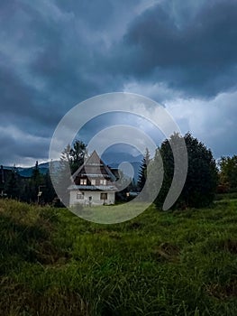 The house is surrounded by trees and mountains, under a dramatic, cloudy sky