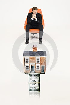 House on top of roll of bills with pensive businessman on chair representing expensive real estate