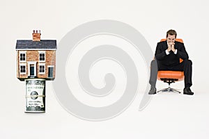 House on top of roll of bills with pensive businessman on chair representing expensive real estate