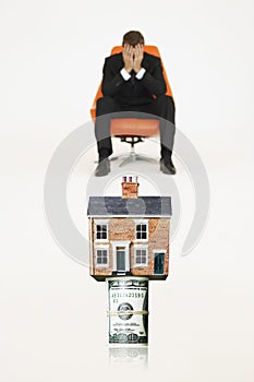House on top of roll of bills with worried businessman on chair representing expensive real estate