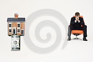 House on top of roll of bills with worried businessman on chair representing expensive real estate