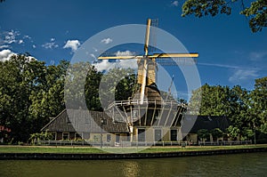 House and wooden yellow windmill next to wide tree-lined canal on the bright light of sunset at Weesp.