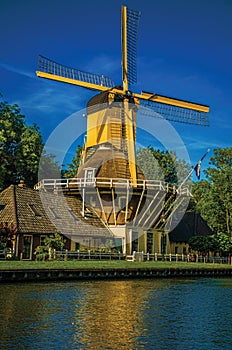 House and wooden yellow windmill next to wide tree-lined canal on the bright light of sunset at Weesp.