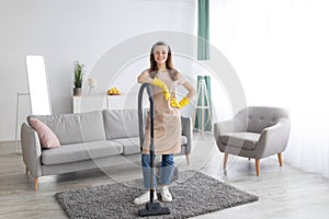 Housecleaning service. Happy young maid with vacuum cleaner standing in middle of tidy living room, full length portrait