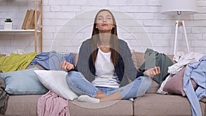 Household and stressless practice. Young calm peaceful woman meditating among scattered garment, sitting on sofa