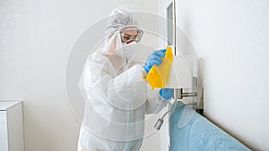Housekeeper or maid cleaning and desinfecting furniture in hotel room. Person wearing protective medical suit doing