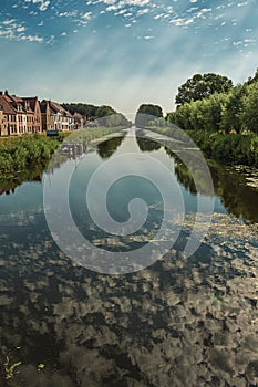 Houses, bushes and grove along canal with sky reflected on water, in the late afternoon and blue sky, near Damme.