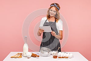 Housewife chef cook confectioner or baker searching new recipe in internet, standing with tablet in hands near table with products