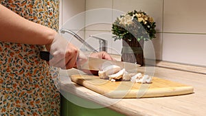 Housewife chopping and slicing fresh chiken meat with steel knife on wooden cutting board on table in domestic kitchen.