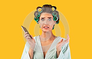 Housewife Holding Phone Looking At Camera In Confusion, Yellow Background