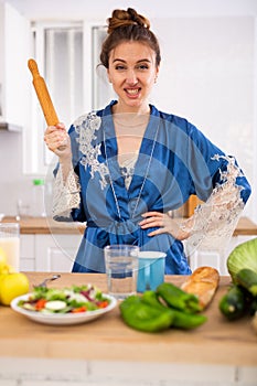 Housewife in a housecoat with a rolling pin in her hands in the kitchen