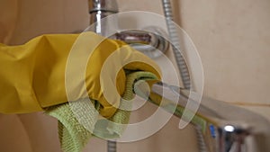 A housewife in protective rubber yellow gloves cleans a stainless steel faucet with a washing gel. Dirty bathroom faucet