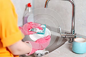 Housework. A housewife in pink rubber gloves is washing a plate. Close up of kitchen sink and hands. View from the shoulders