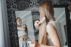 How take care of long hair. Back view of young natural blonde woman in white home clothes combing her hair and looking